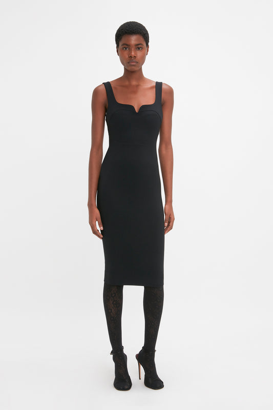 A person stands facing forward, showcasing a Sleeveless Fitted T-Shirt Dress In Black by Victoria Beckham, paired with black lace leggings and black heeled shoes. The backdrop is plain white.