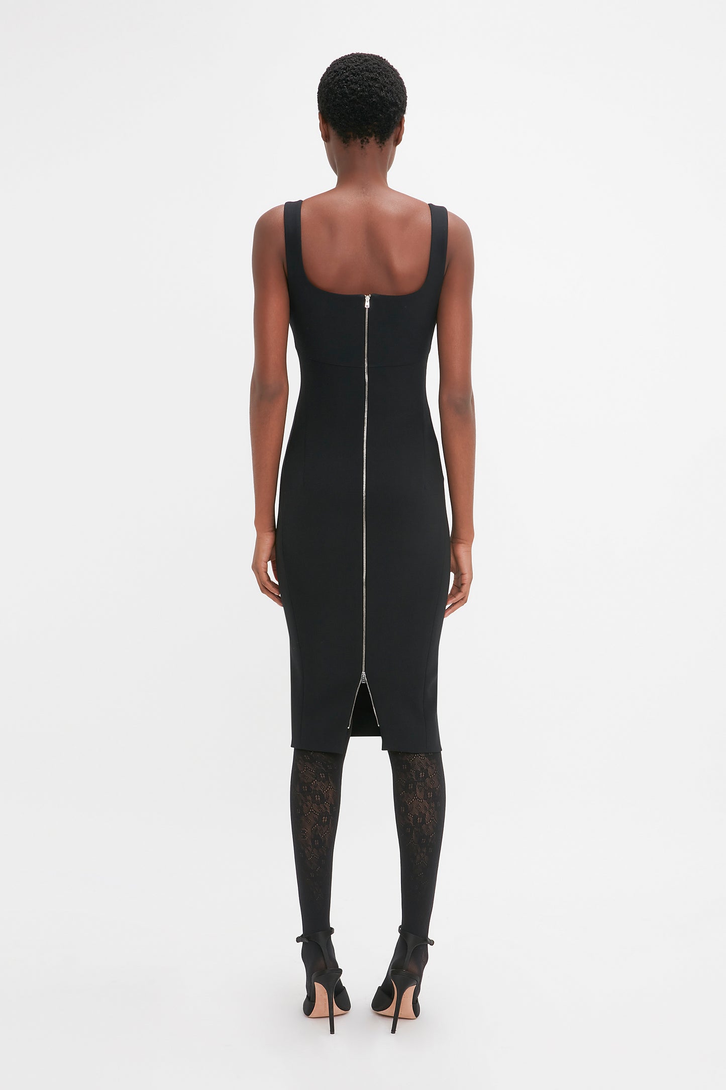 A person stands facing away, wearing a Sleeveless Fitted T-Shirt Dress In Black by Victoria Beckham with a visible back zipper, black lace-patterned tights, and black high heels. Hair is short and curly. The background is plain white.