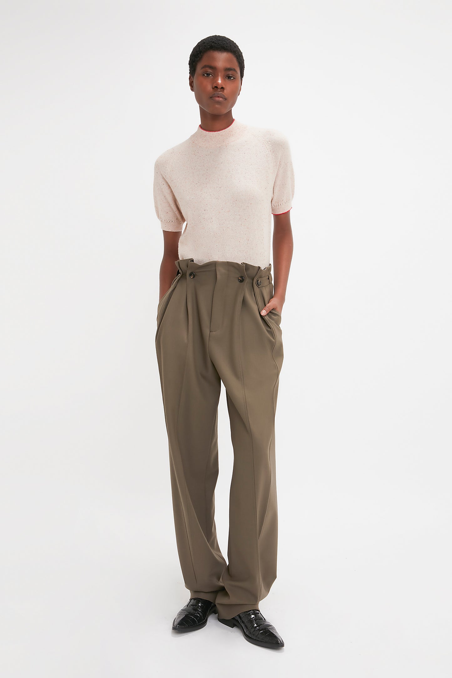 A person wearing a light-colored Short Sleeve Top In Nougat by Victoria Beckham and high-waisted, olive green pants with hands in pockets, standing against a plain white background. The lightweight cashmere knit top adds a soft touch to the effortlessly chic outfit.