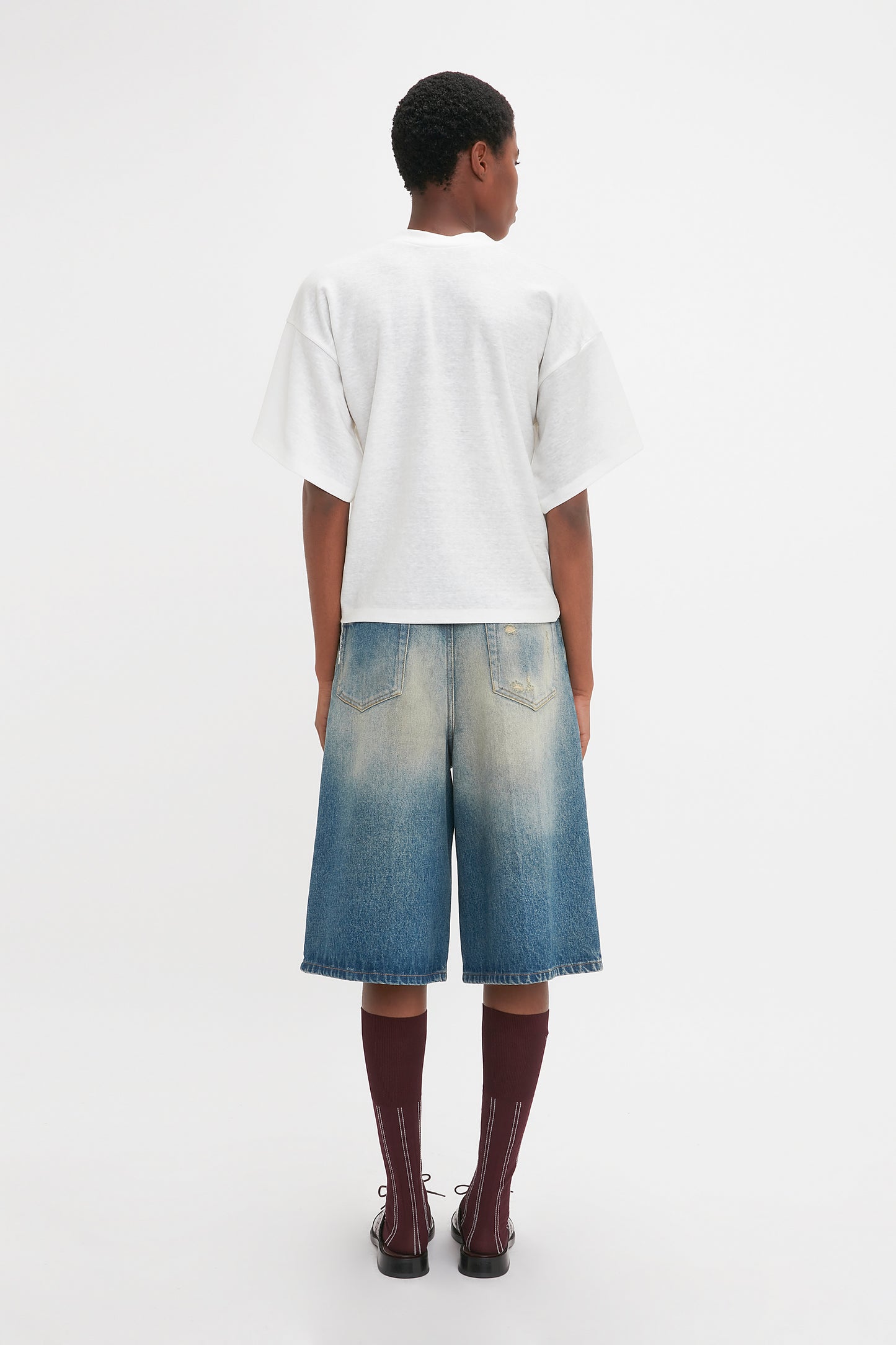Person standing with their back to the camera. They are wearing a plain white t-shirt, Victoria Beckham Oversized Bermuda Short In Antique Indigo Wash, maroon socks, and black shoes. The background is plain white.