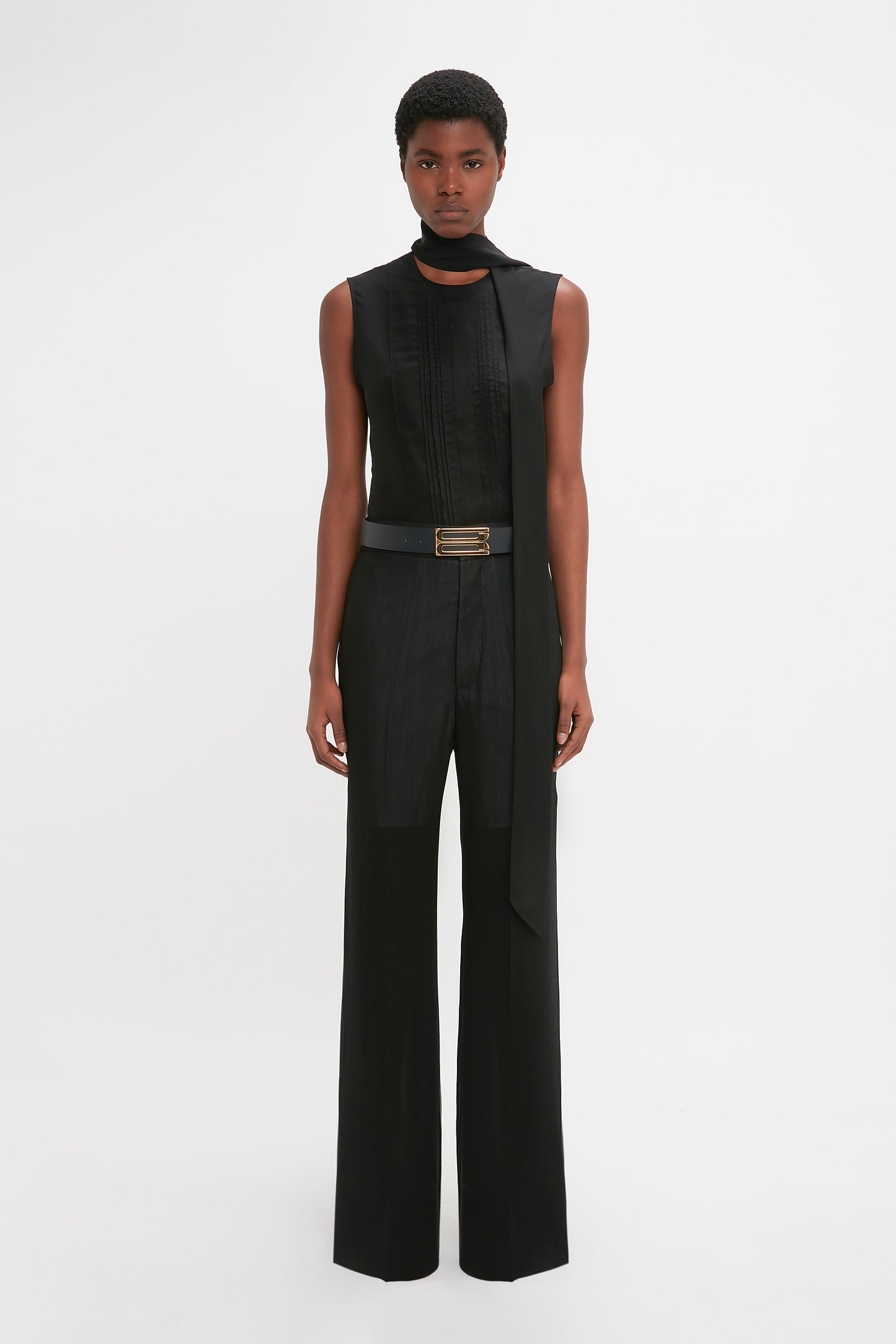 A black woman stands facing the camera, wearing a modern Victoria Beckham sleeveless top and high-waisted, Waistband Detail Straight Leg Trouser In Black with a belt.