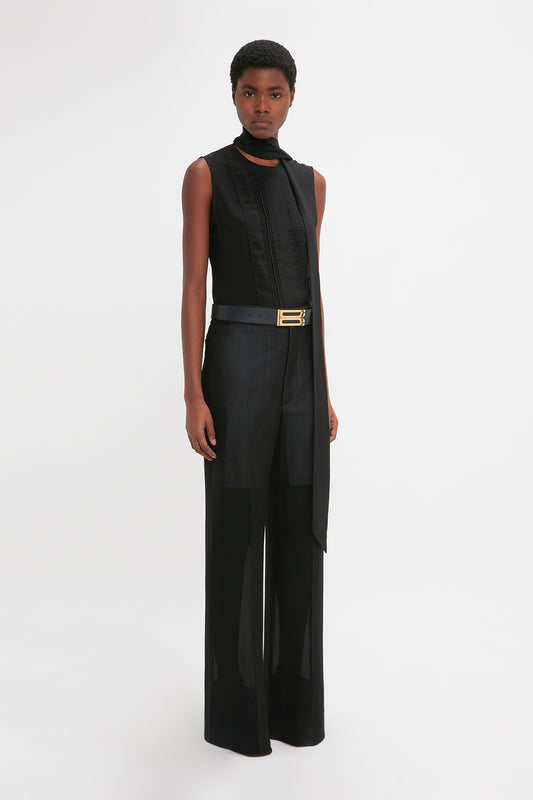 A woman wearing Victoria Beckham's Waistband Detail Straight Leg Trouser In Black and a sleek black sleeveless turtleneck, with a long, draped side panel and a golden belt, standing against a white background.