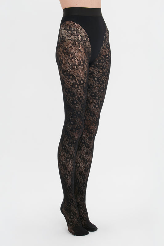 Woman wearing black Victoria Beckham Monogram Lace Tights with seamless and sag-free construction on a white background, standing sideways.