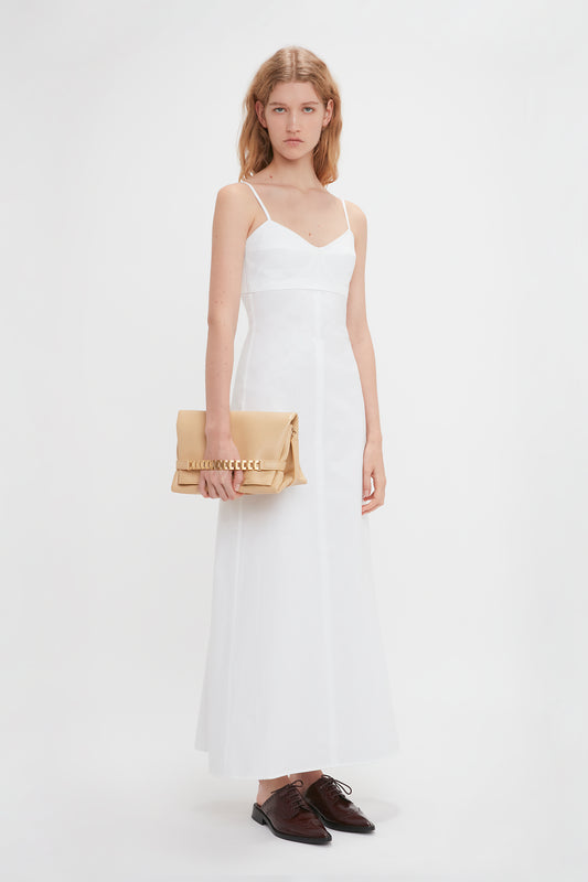 A woman in a white sleeveless dress stands holding a Victoria Beckham sesame leather chain pouch with strap, with brown shoes beside her on a plain white background.