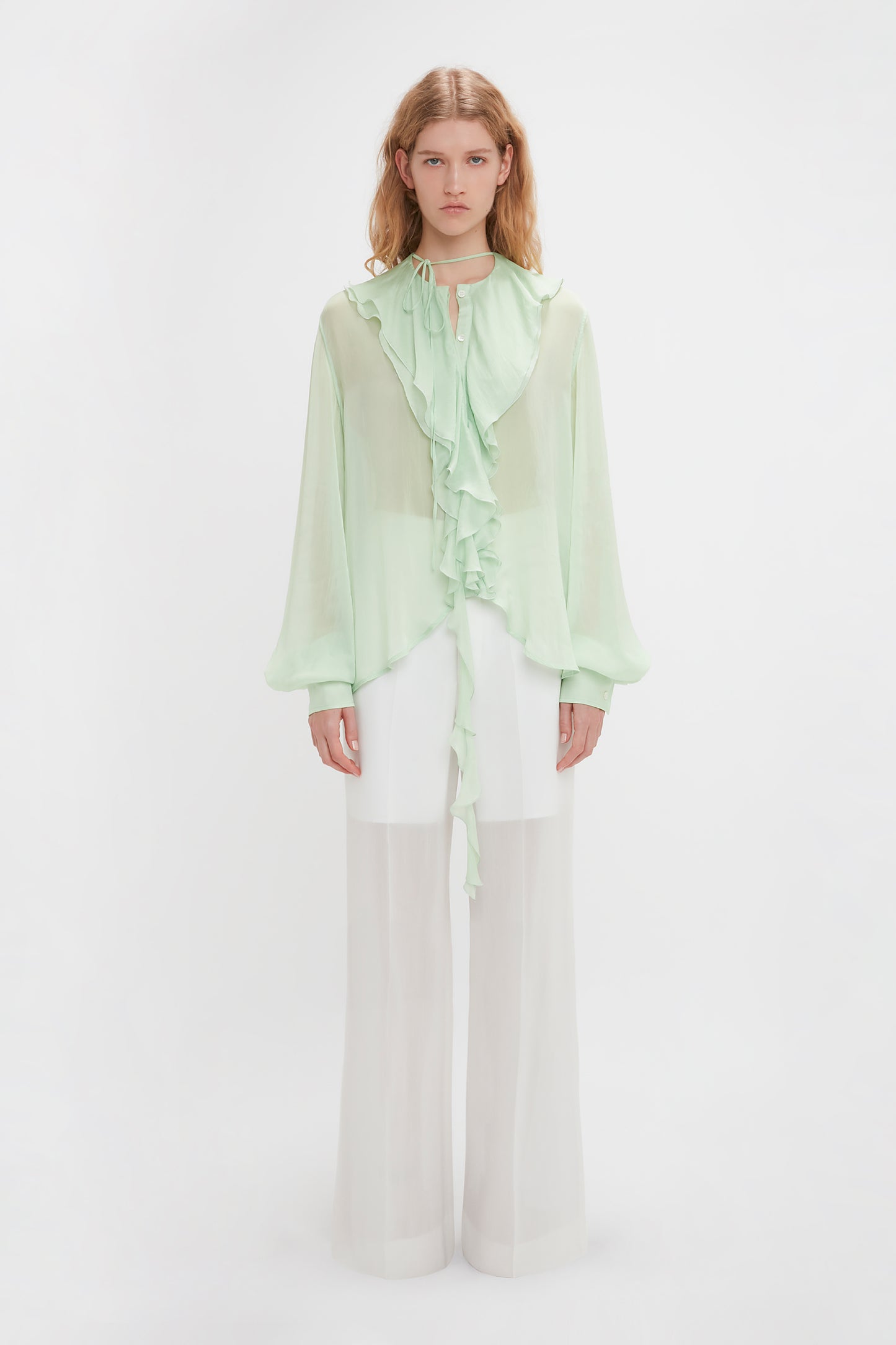 A person stands against a plain white background, adorned in a Romantic Blouse In Jade by Victoria Beckham with diaphanous blouson sleeves, paired with white wide-legged sheer trousers.