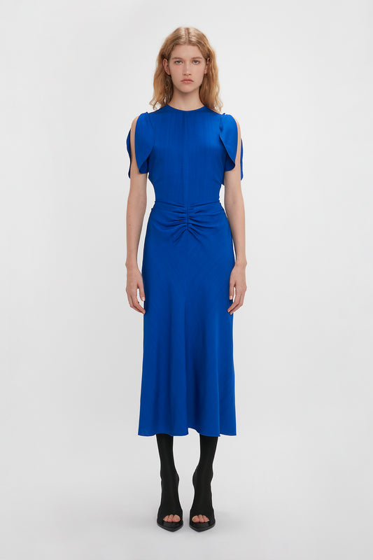 A woman in a Victoria Beckham Gathered Waist Midi Dress In Palace Blue with cap sleeves and a knotted detail at the waist, standing against a white background.