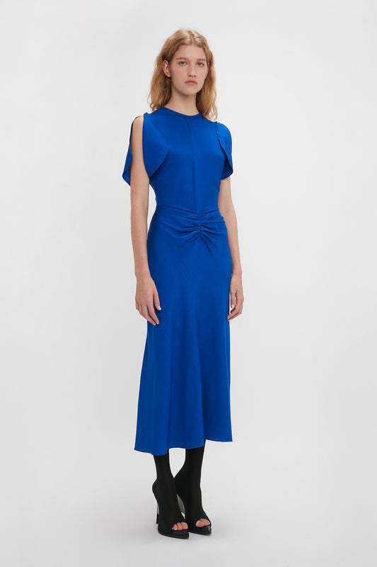A woman in a Victoria Beckham gathered waist midi dress in palace blue with cap sleeves and a front twist detail, paired with black open-toe heels, standing against a plain white background.