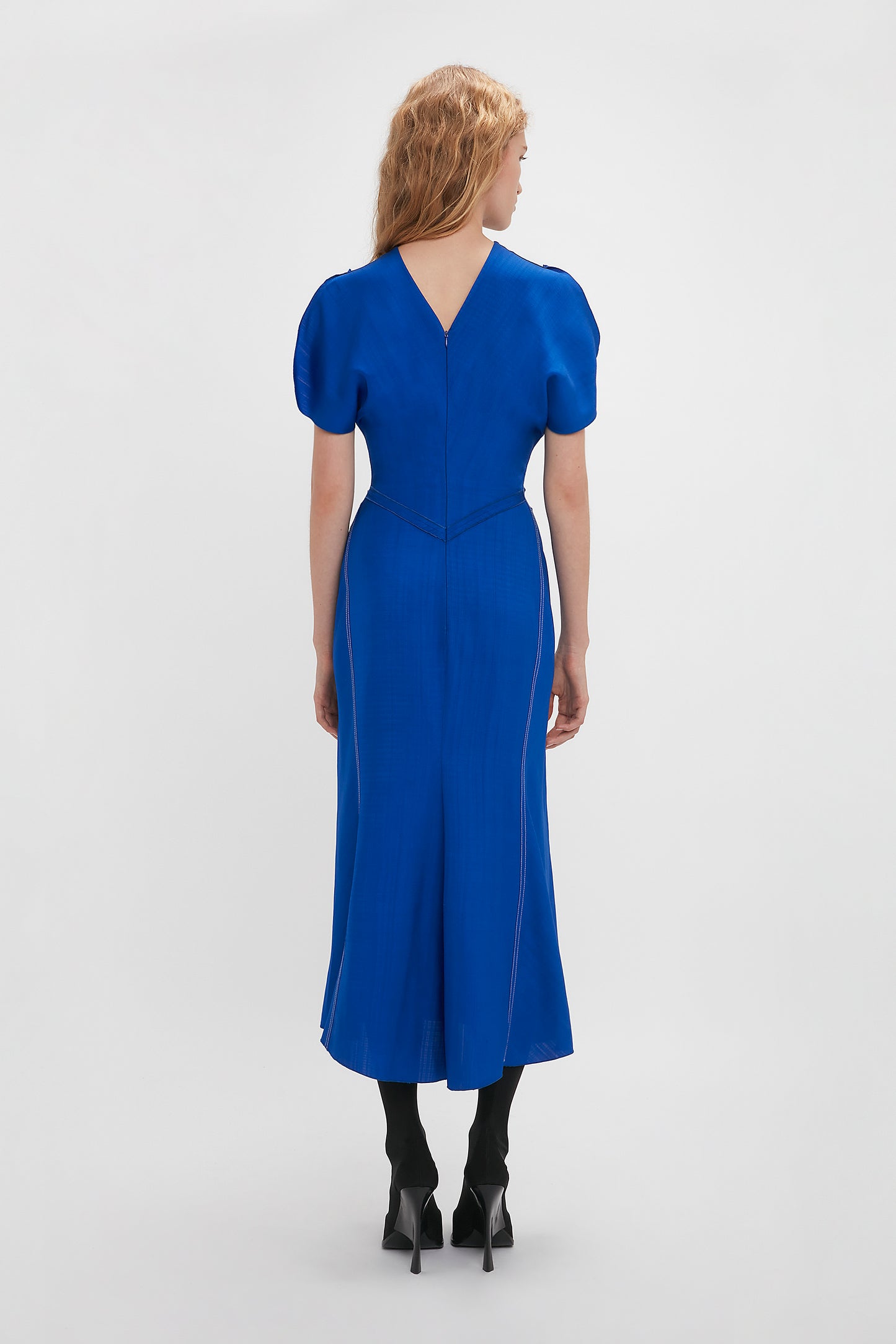 Woman in a Victoria Beckham Palace Blue Gathered Waist Midi Dress and black heels standing with her back to the camera against a white background.