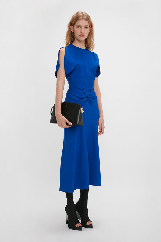 Woman in a Victoria Beckham Gathered Waist Midi Dress In Palace Blue and black heels carrying a black clutch bag, standing against a white background.
