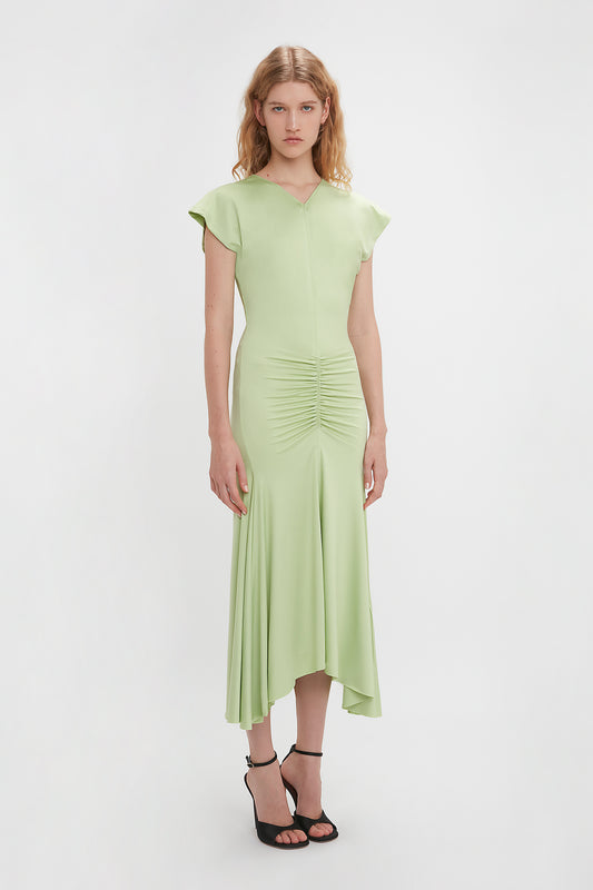 A woman models a Victoria Beckham Sleeveless Ruched Jersey Dress In Pistachio with cap sleeves and ruched detailing, paired with black strappy heels, against a white background.