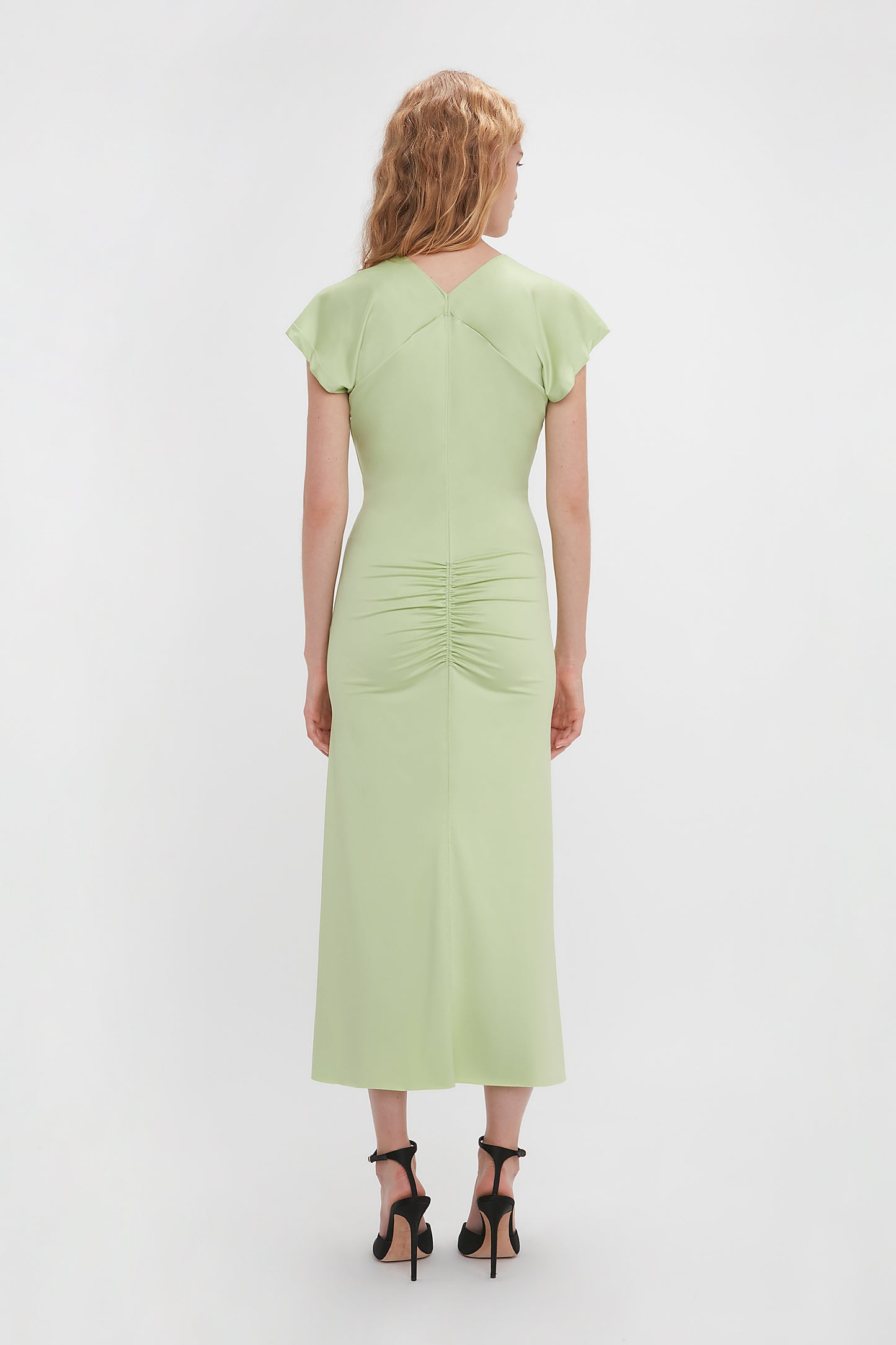 A person with long hair is standing facing away, wearing a Victoria Beckham Sleeveless Rouched Jersey Dress In Pistachio that highlights their body-skimming silhouette and black high-heeled shoes against a plain white background.