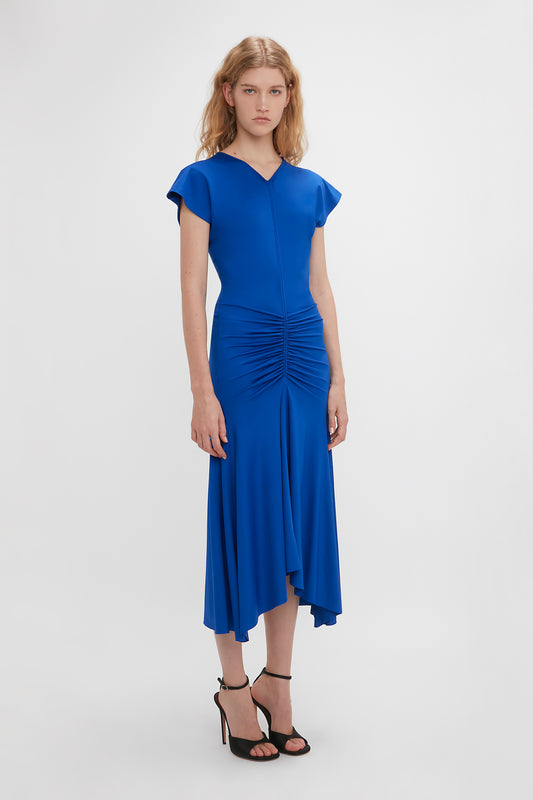 A woman stands against a white background, wearing a bright Victoria Beckham royal blue knee-length dress with ruched detailing and short sleeves, paired with black high-heeled sandals.