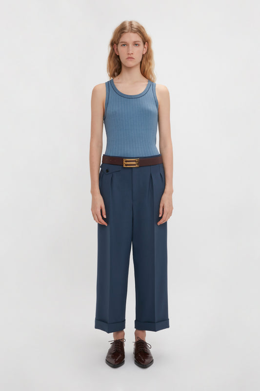 A woman standing against a white background, wearing a blue fine-knit tank top, Victoria Beckham's Wide Leg Cropped Trousers in Heritage Blue, and brown shoes.