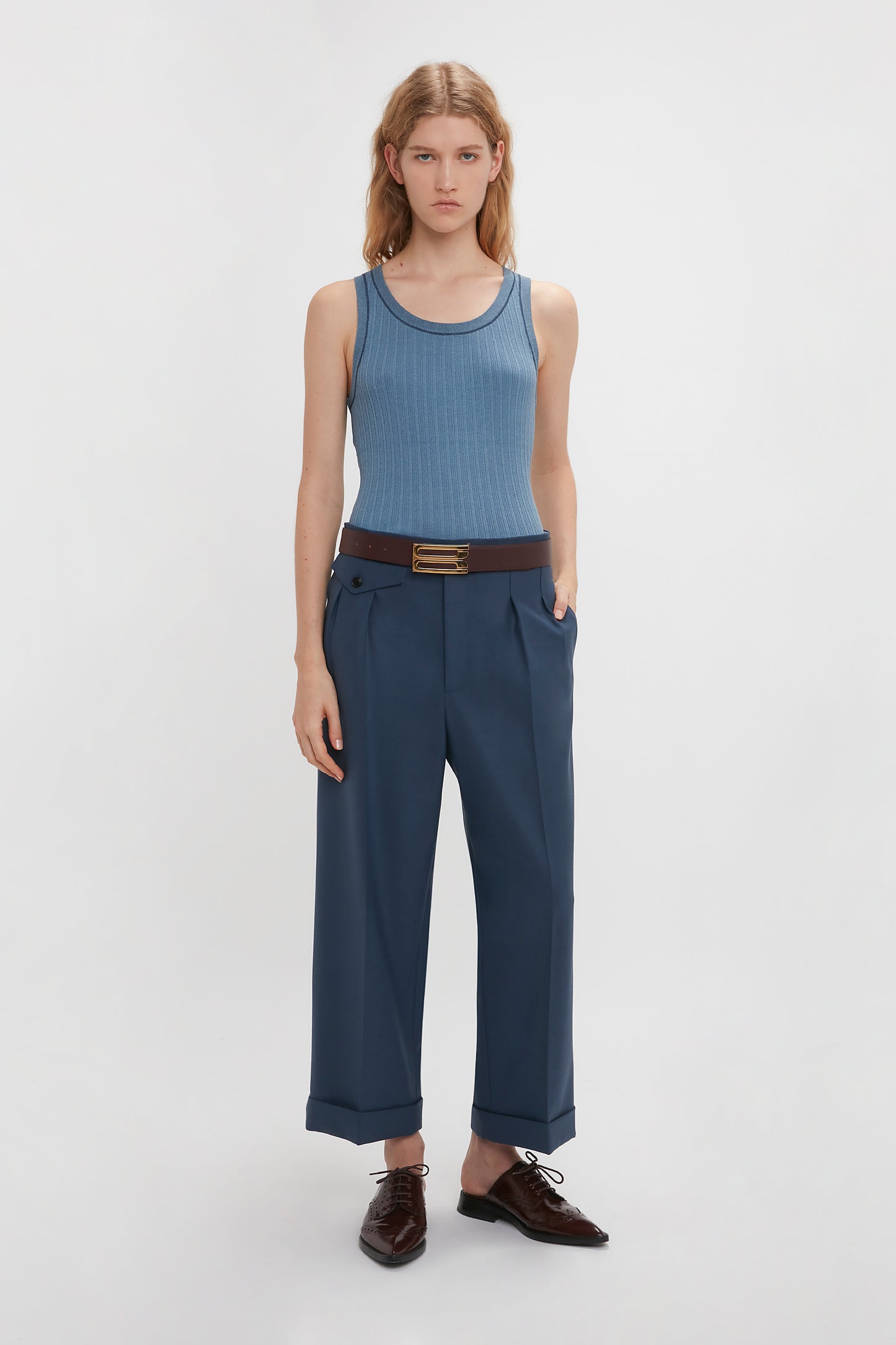 Person standing against a white background, wearing a Victoria Beckham Fine Knit Micro Stripe Tank In Heritage Blue, dark blue trousers with a black belt, and dark brown shoes. Their hands are in their pockets.