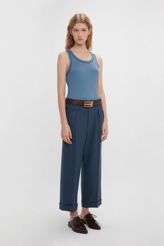 A woman in a blue fine knit tank and Victoria Beckham navy wide-leg cropped trousers with a brown belt, standing against a white background.