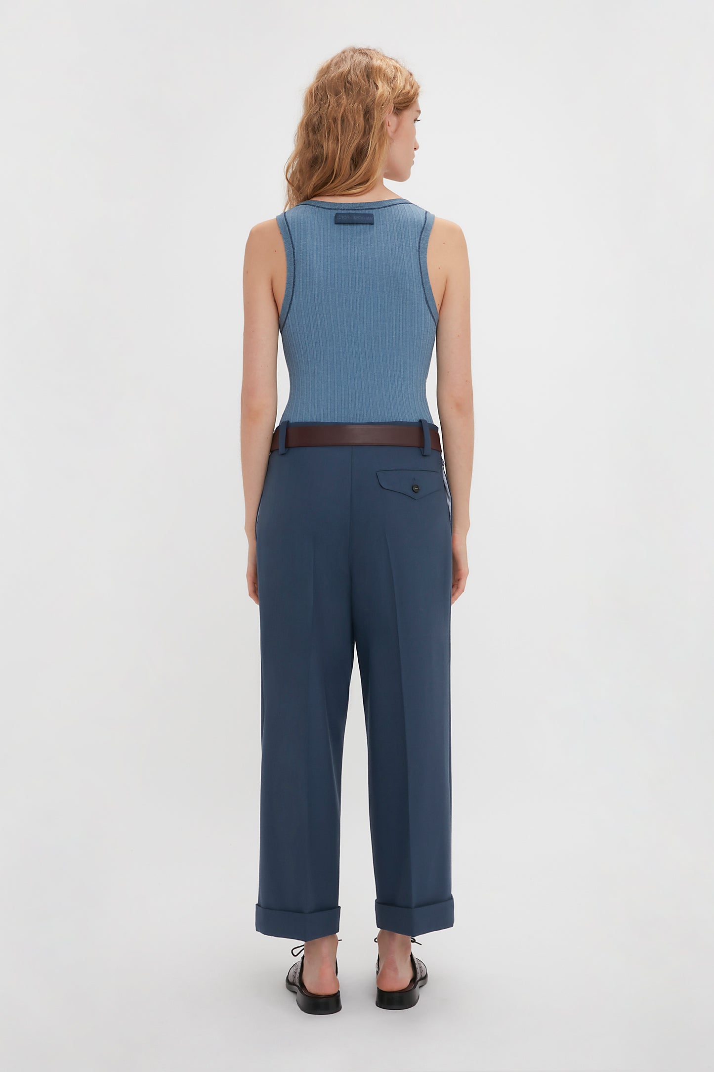 A woman stands facing away from the camera, wearing a blue fine knit tank and Victoria Beckham wide leg cropped trousers in heritage blue in a plain white studio setting.