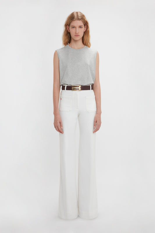A woman in a Victoria Beckham grey marl sleeveless t-shirt and white flared jeans, accessorized with a black belt with a gold buckle, standing against a plain white background.