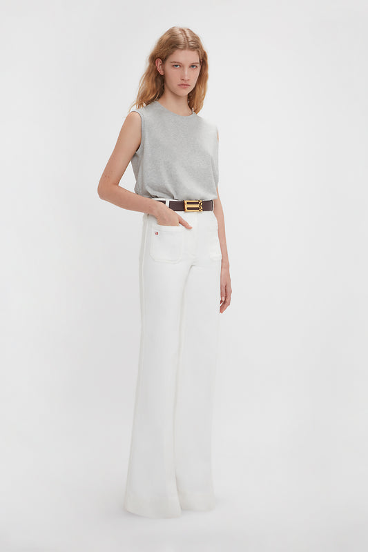 A woman models high-waisted white trousers paired with a Victoria Beckham sleeveless t-shirt in grey marl, accessorized with a black belt with a gold buckle, standing against a plain white background.