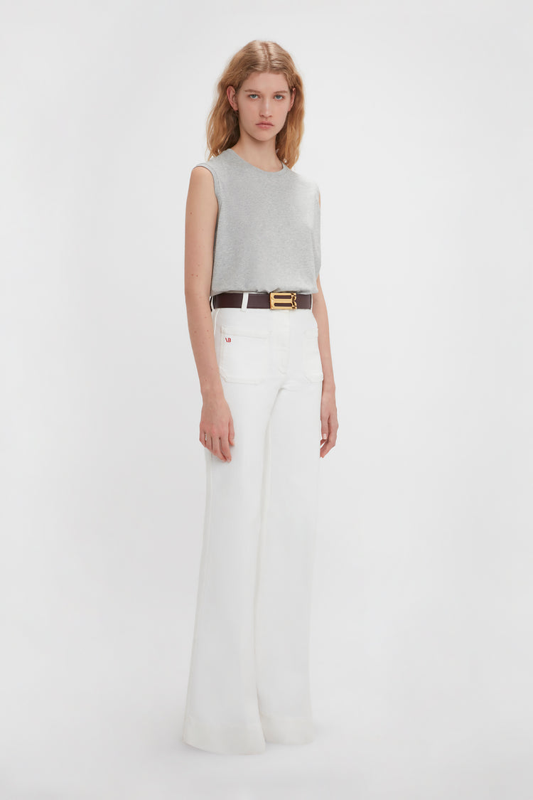 A woman in a Victoria Beckham grey marl sleeveless t-shirt and white wide-legged pants standing against a plain white background. She wears a black belt with a gold buckle.