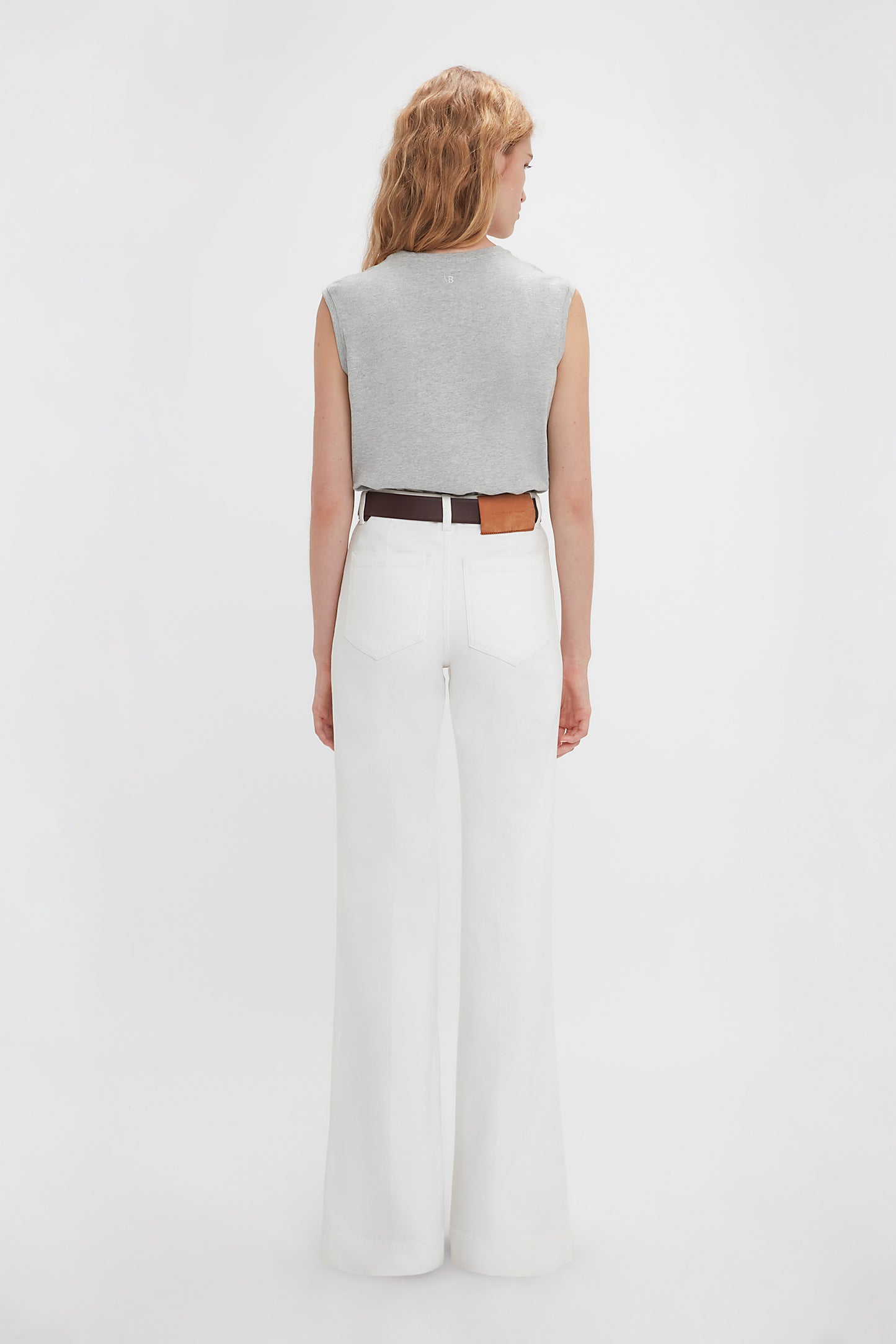 Woman viewed from behind wearing white wide-leg pants and a Victoria Beckham sleeveless t-shirt in grey marl, accessorized with a brown belt.