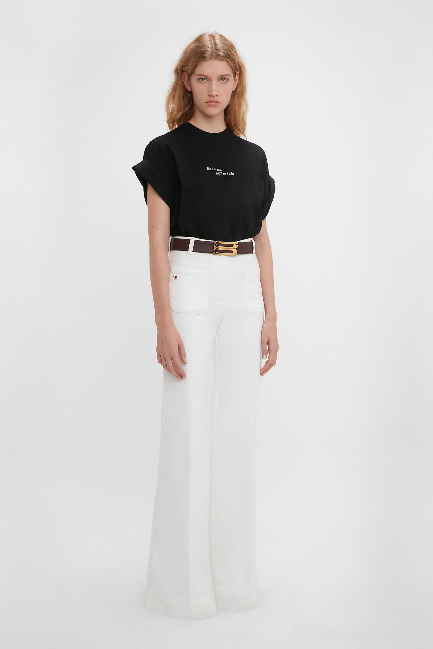 A woman wearing a black 'Do As I Say, Not As I Do' Slogan T-shirt by Victoria Beckham and white high-waisted flared jeans with a slim brown belt, standing against a plain white background.