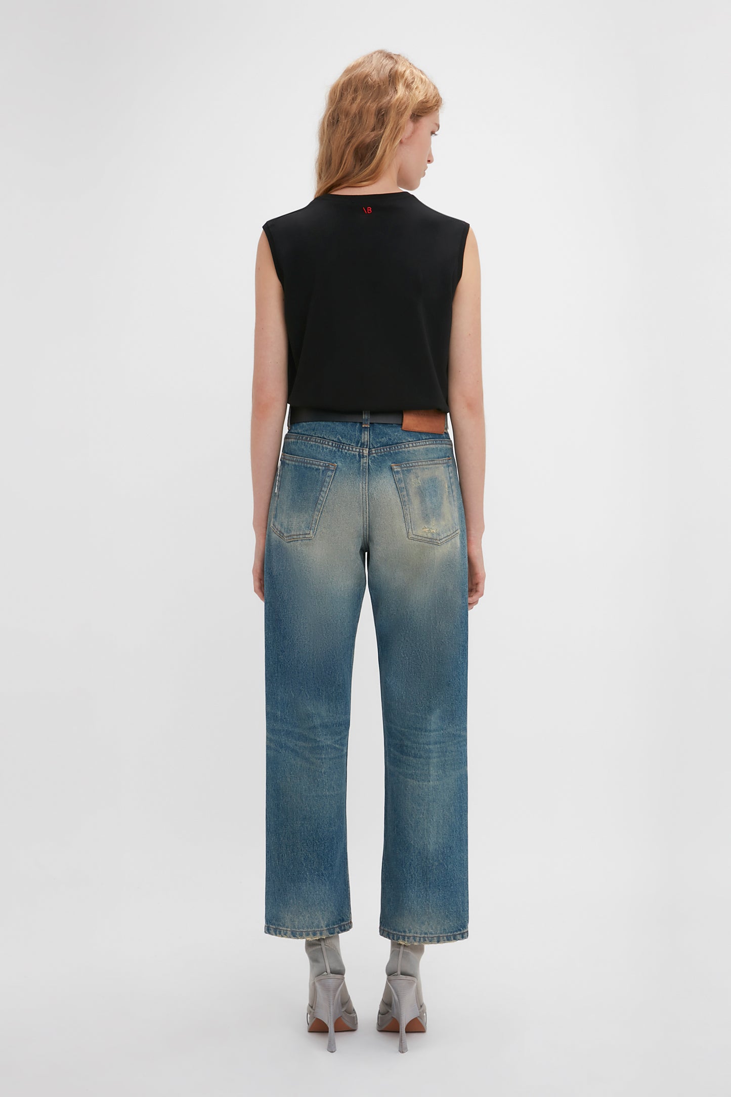 Woman standing with her back to the camera, wearing a black sleeveless top and blue Victoria Beckham Relaxed Straight Leg Jeans in Antique Indigo Wash, against a white background.