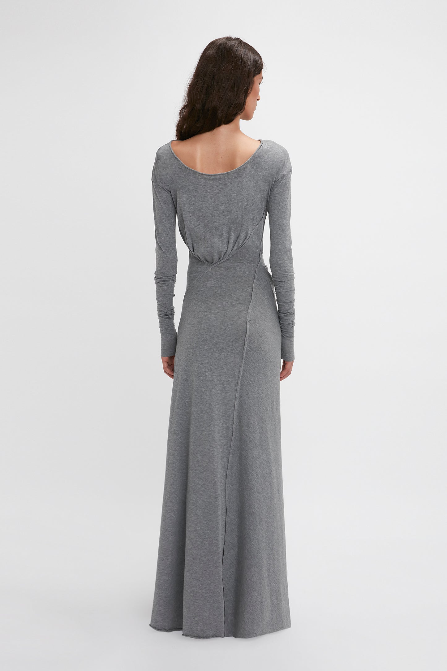 A person with long, dark hair is standing with their back to the camera, wearing a Long Sleeve Circle Neck Dress In Grey Marl by Victoria Beckham crafted from a super-soft cotton-jersey blend. The floor-length grey garment exudes an elegant touch of contemporary eveningwear.