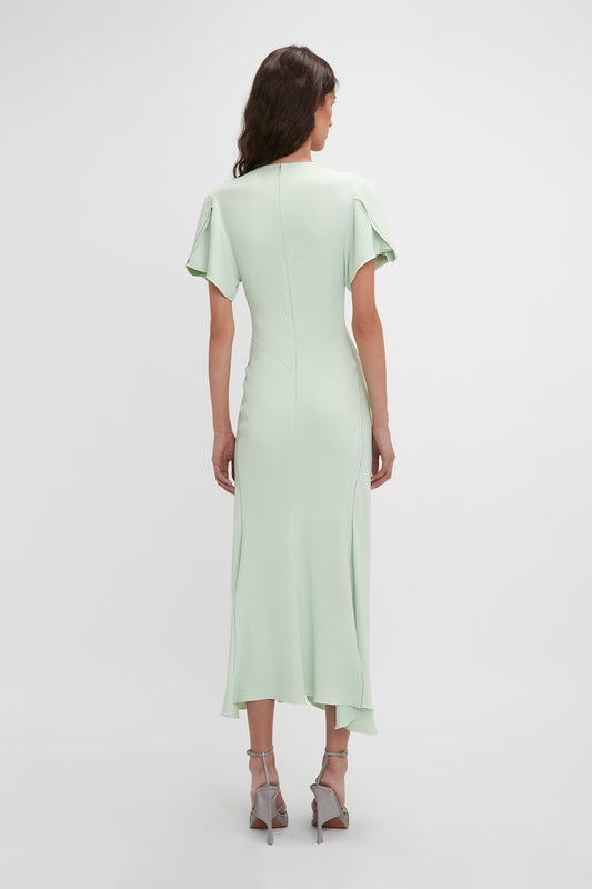 A woman in a light green Victoria Beckham Gathered V-Neck Midi Dress In Jade with flutter sleeves, viewed from the back, standing against a plain white background.