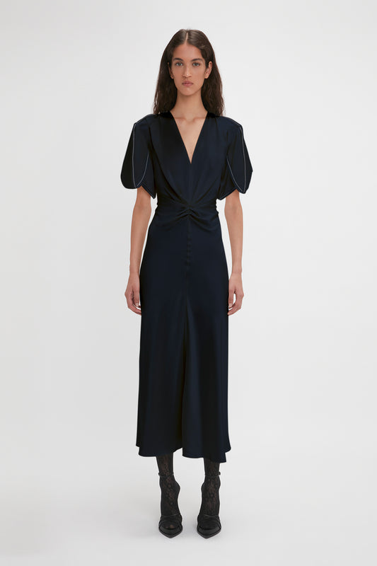A woman wearing a dark blue Exclusive Gathered V-Neck Midi Dress In Navy with puff sleeves and waist-defining pleat detail, paired with black lace-up boots, standing against a white background by Victoria Beckham.