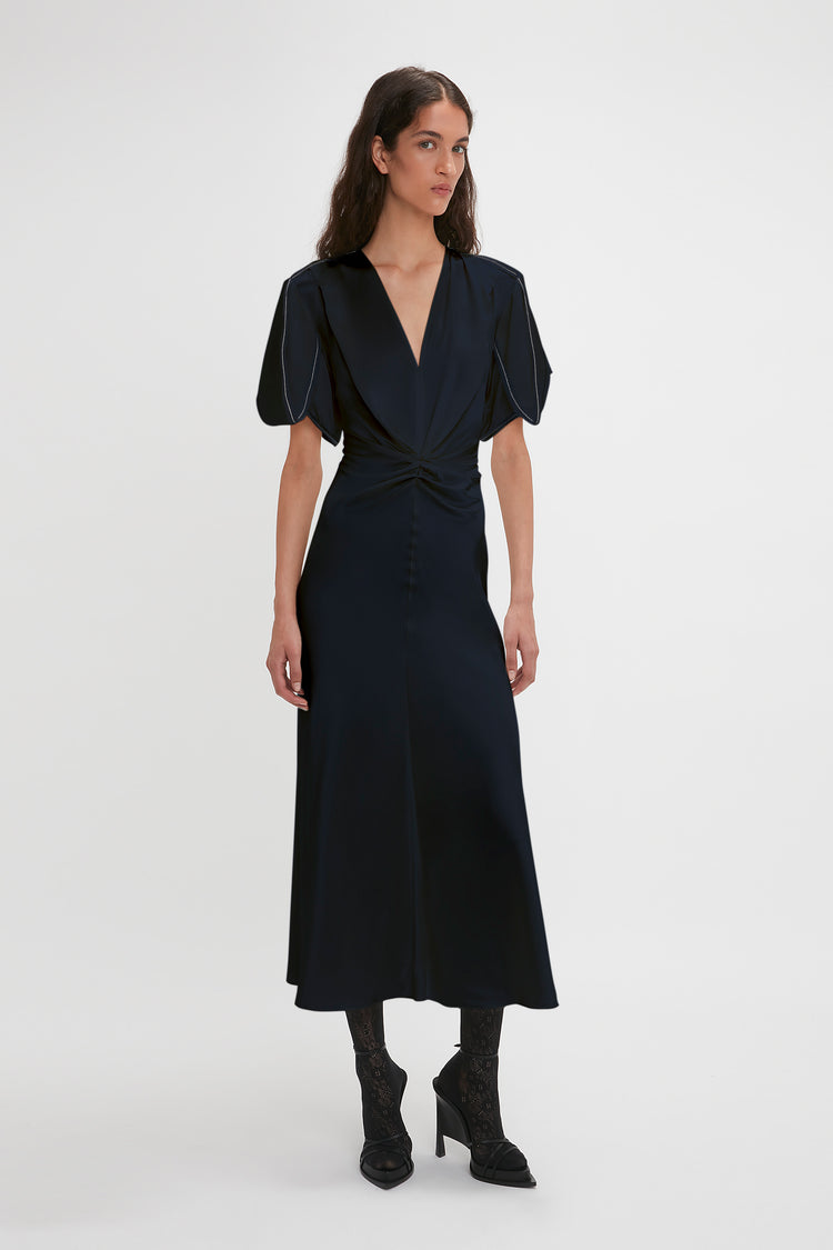 A woman in a Victoria Beckham Exclusive Gathered V-Neck Midi Dress In Navy with waist-defining pleat detail and puff sleeves, paired with black lace-up boots, standing against a white background.