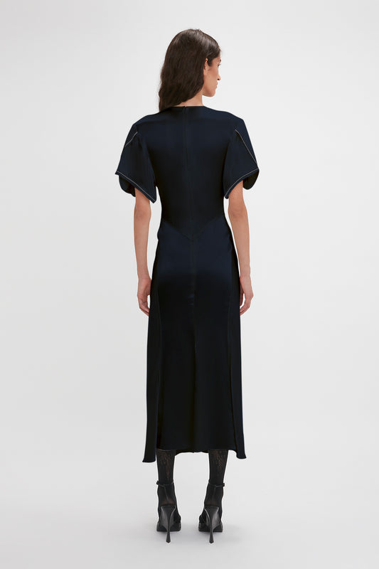 A woman standing with her back to the camera, wearing a dark Victoria Beckham Exclusive Gathered V-Neck Midi Dress In Navy with short sleeves and black heeled boots on a plain background.