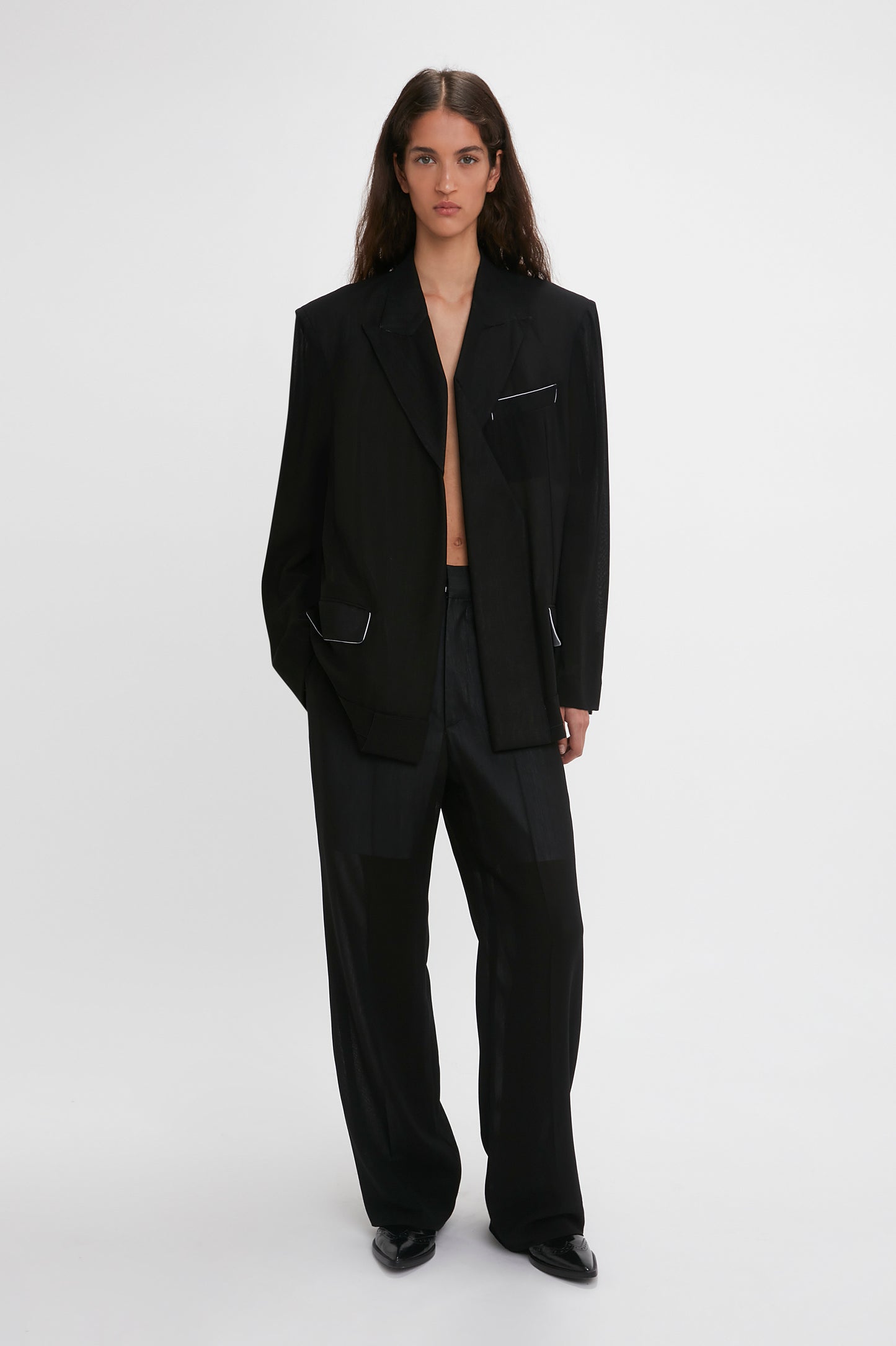 A woman models a Victoria Beckham black wool Fold Detail Tailored Jacket with waistband detail straight leg trousers, standing against a plain white background.