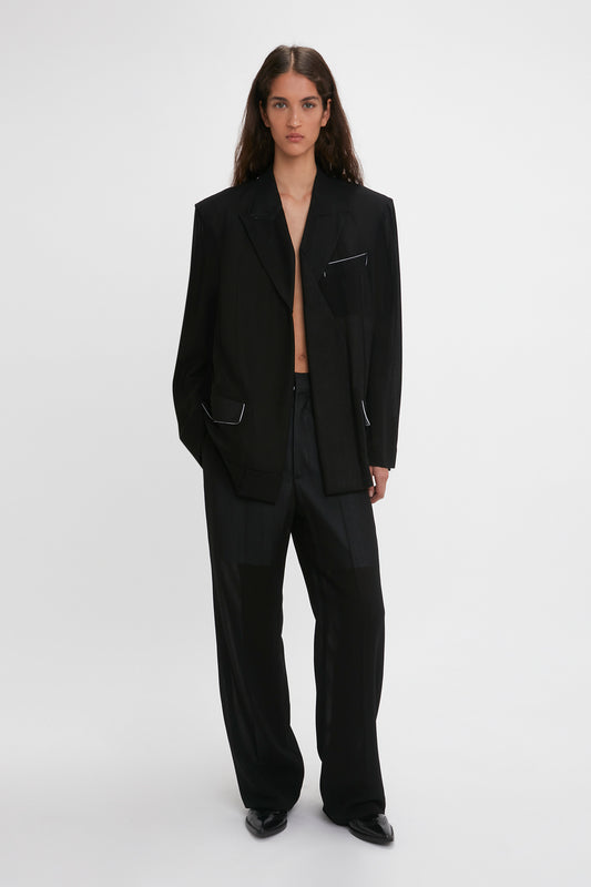 A woman models a Victoria Beckham black wool Fold Detail Tailored Jacket with waistband detail straight leg trousers, standing against a plain white background.