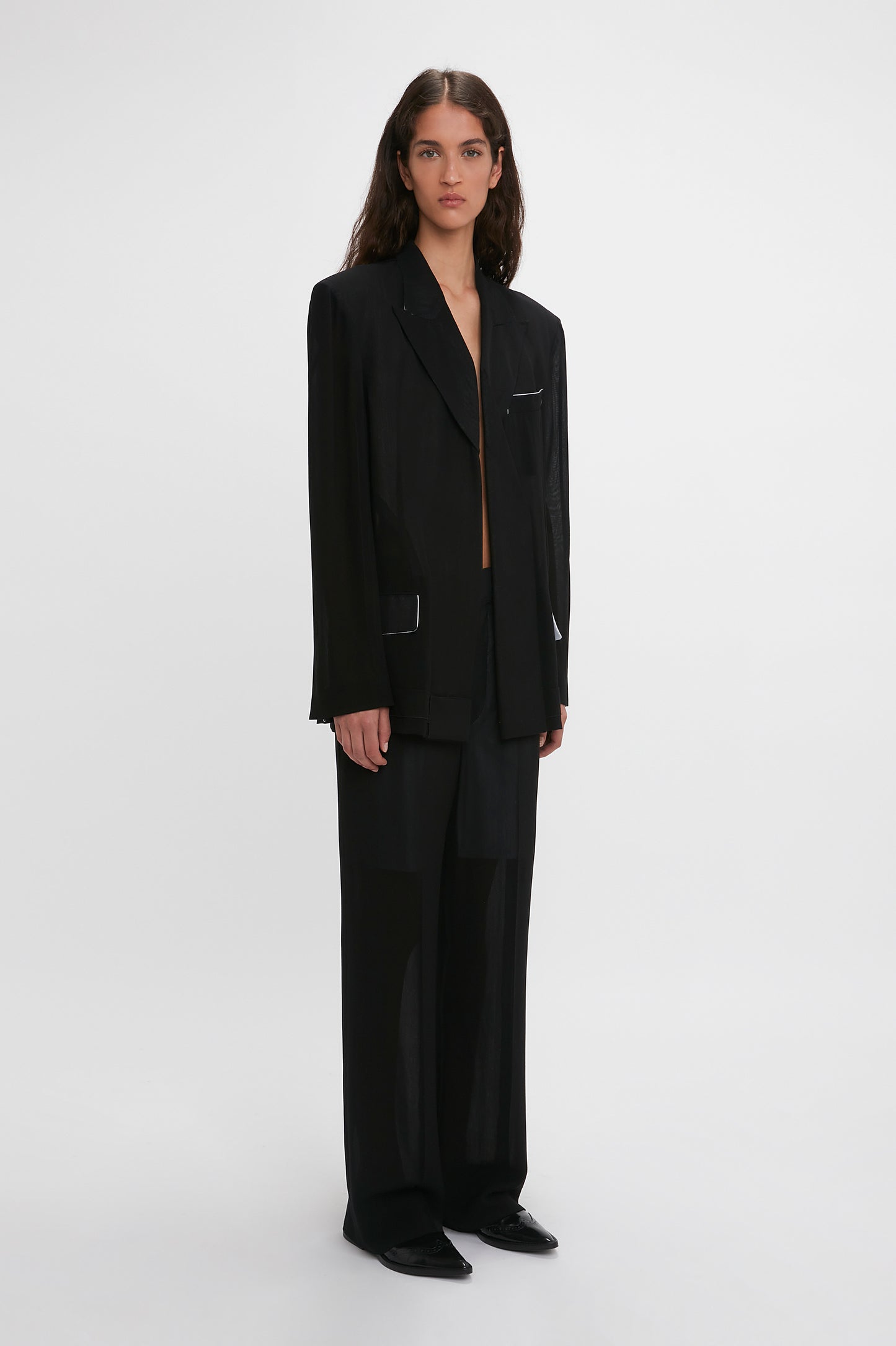 A woman in a Victoria Beckham Fold Detail Tailored Jacket In Black and wool trousers, standing against a white background.