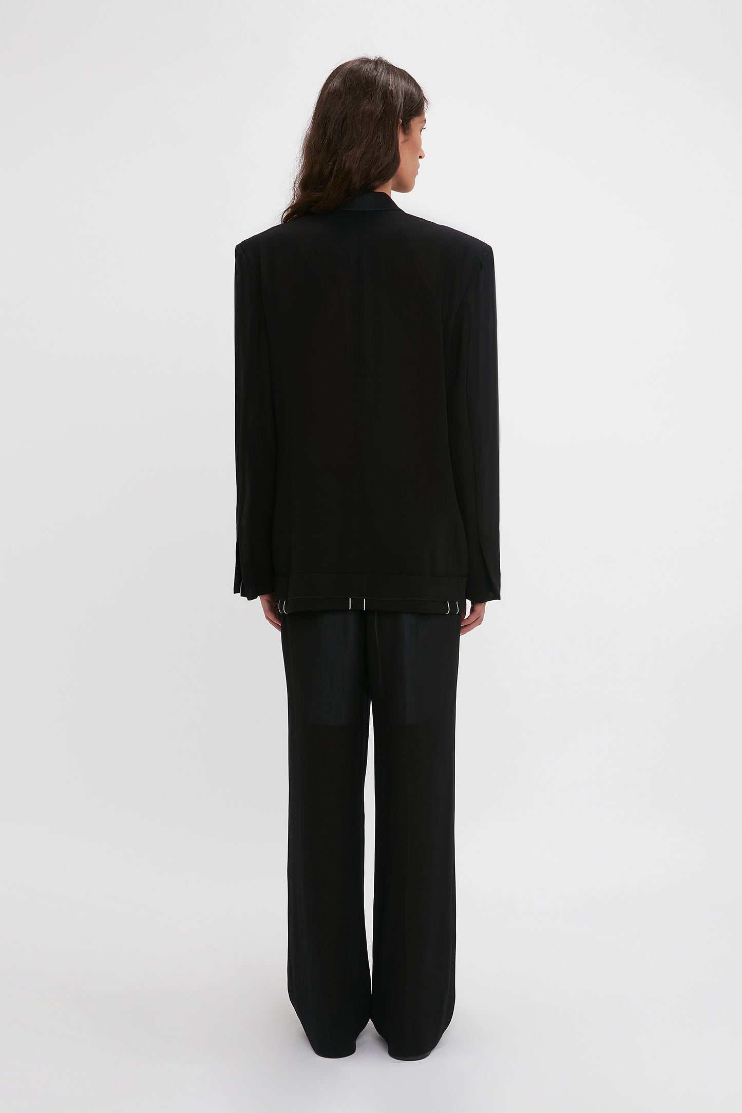 Woman in an elegant Victoria Beckham black pantsuit with waistband detail straight leg trousers, standing with her back to the camera against a white background.