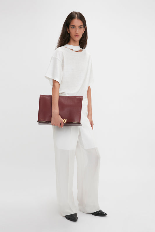 A woman in a white t-shirt and flowing white pants with Victoria Beckham Waistband Detail Straight Leg Trousers holds a maroon handbag, standing against a plain background.