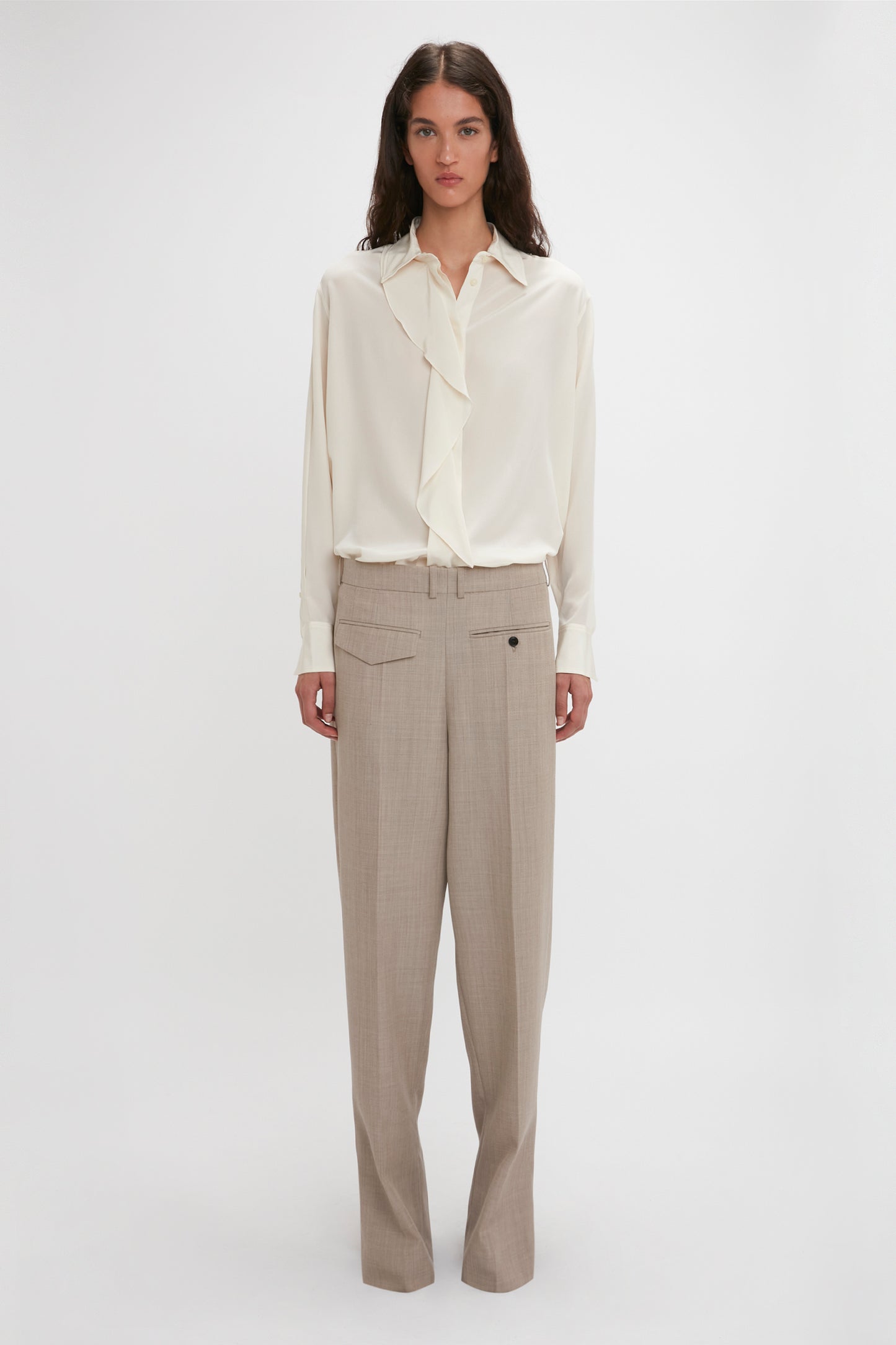 A person stands against a plain background wearing an Asymmetric Ruffle Blouse In Ivory by Victoria Beckham, featuring voluminous sleeves, and beige trousers.