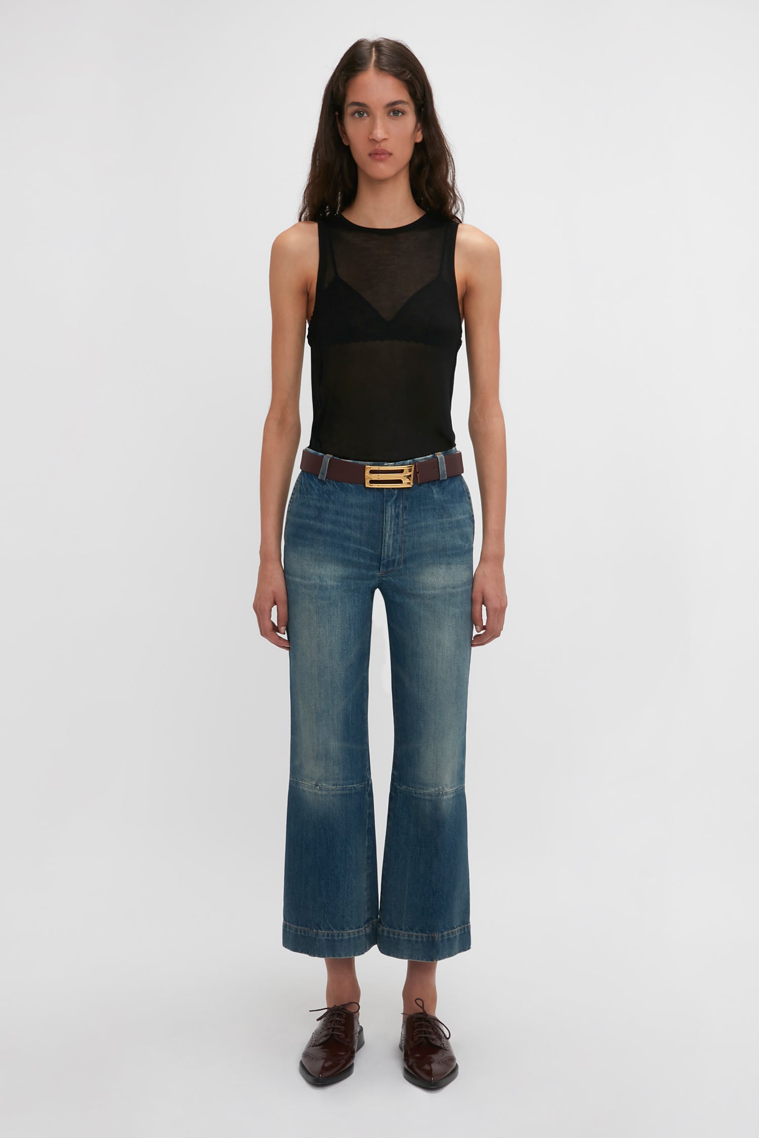 A woman stands facing forward, wearing a Lightweight Tank Top In Black by Victoria Beckham, blue wide-legged jeans, a brown belt, and dark brown shoes against a plain white background.