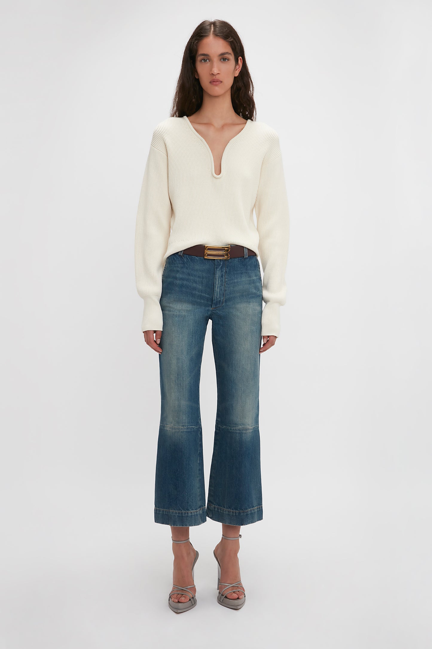 A woman in a white v-neck sweater and Victoria Beckham Cropped Kick Jean In Indigrey Wash stands on a plain background, facing the camera.