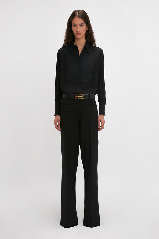 A woman stands against a white backdrop wearing a Victoria Beckham Reverse Front Trouser In Black with contemporary silhouette and a belted waist.