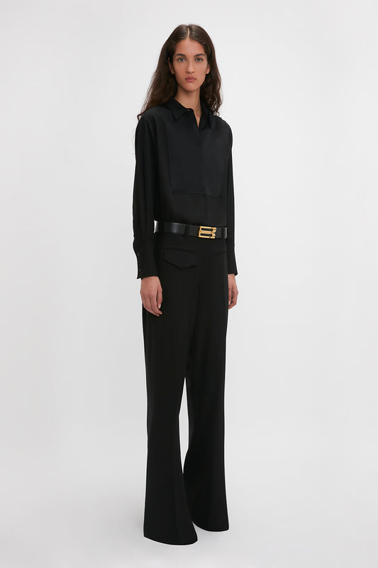A woman in a black blouse and Victoria Beckham's Reverse Front Trouser In Black with a wide belt, posing against a white background.