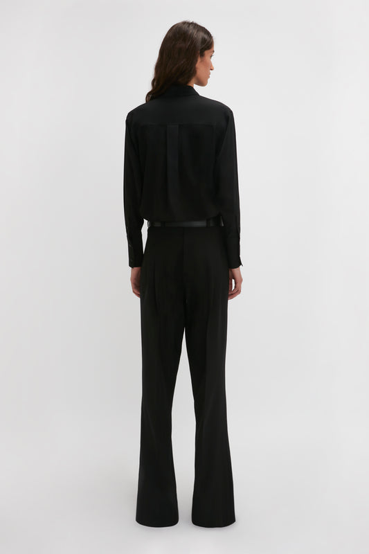 A woman in a black pantsuit stands facing away, showcasing the tailored back design of the Victoria Beckham Reverse Front Trouser In Black on a plain light background, highlighting the contemporary silhouette.