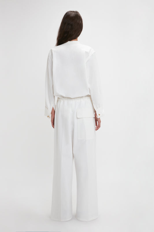 A woman standing with her back to the camera, wearing an elegant white cotton-canvas pantsuit with a Victoria Beckham Drawstring Pyjama Trouser In Washed White with an adjustable drawstring waistband and a large pocket on the right side.