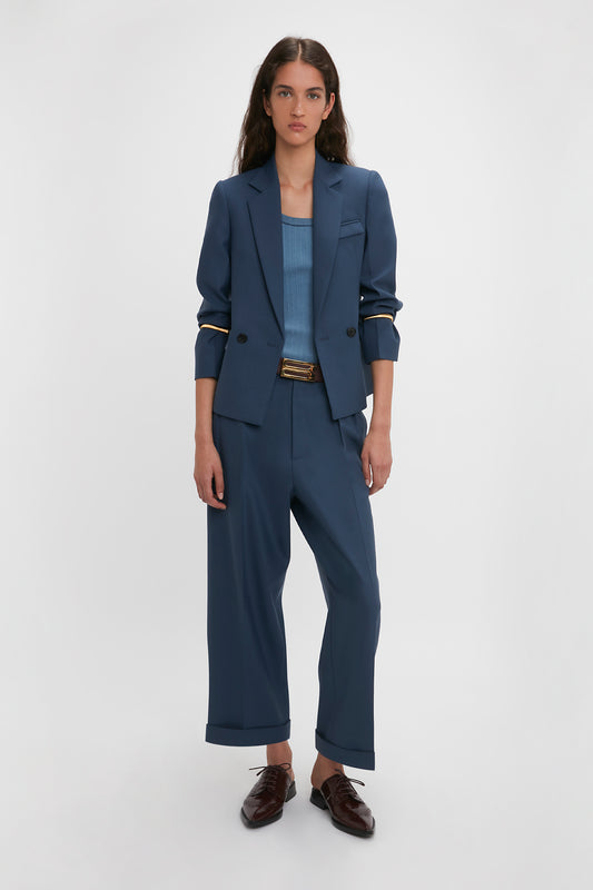 A woman wearing a Victoria Beckham Shrunken Double Breasted Jacket In Heritage Blue with cropped pants and a belt, poised against a plain white background. She complements her outfit with brown loafers.