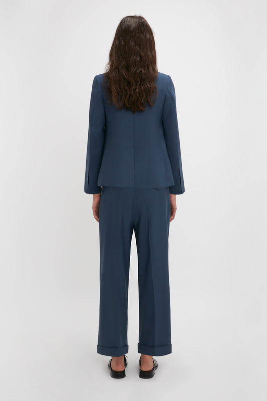 Rear view of a woman wearing a Victoria Beckham Heritage Blue shrunken double-breasted jacket and black shoes, standing against a plain white background.