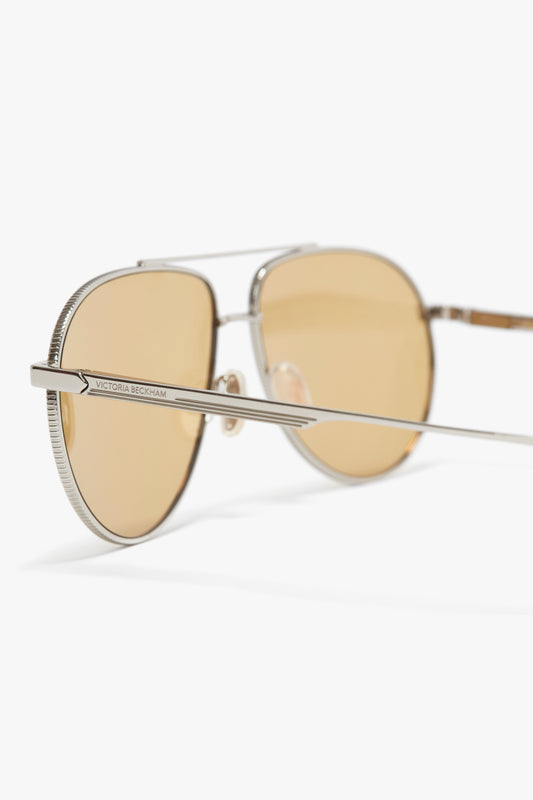 A close-up of Victoria Beckham V Metal Pilot sunglasses in silver-brown with thin metal frames, adjustable nose pads, and amber-tinted lenses, isolated on a white background.