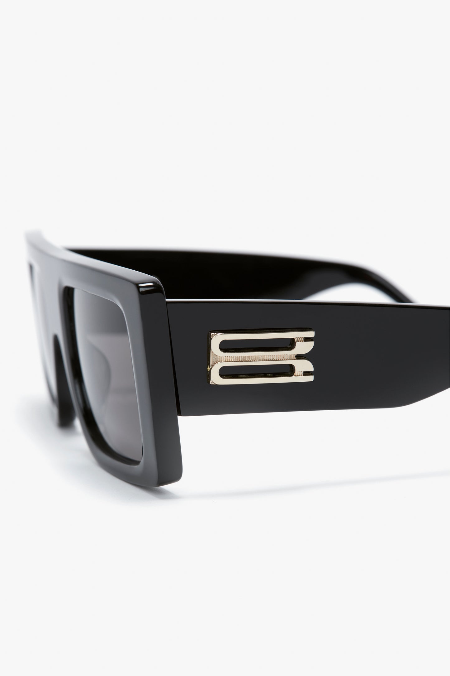 A close-up image of Victoria Beckham Oversized Frame Sunglasses In Black featuring black rectangular lenses and a gold decorative emblem on the side of the bold, oversized frame.