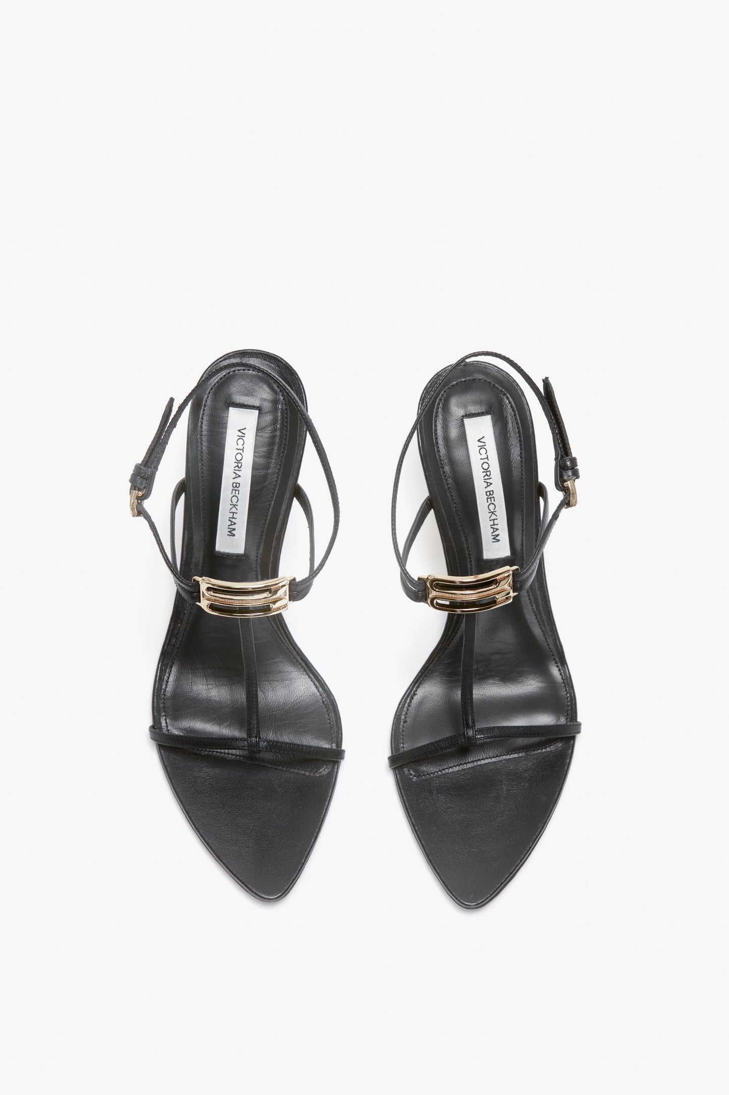 A pair of black, open-toe high-heel sandals with thin ankle straps and gold buckle details, featuring a V-shaped sculptural heel and crafted from supple nappa leather, viewed from above—Frame Detail Sandal In Black Leather by Victoria Beckham.