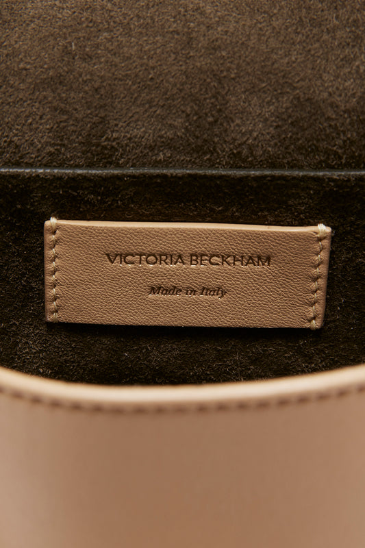 Close-up of a "Victoria Beckham Mini Pouch With Long Strap In Sesame Leather" label on a brown leather crossbody shoulder bag with stitching detail.