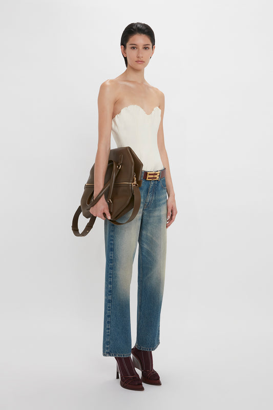 A woman in a Victoria Beckham Antique White corset top, blue jeans with a black belt, maroon heels, and holding a brown handbag stands against a white background.